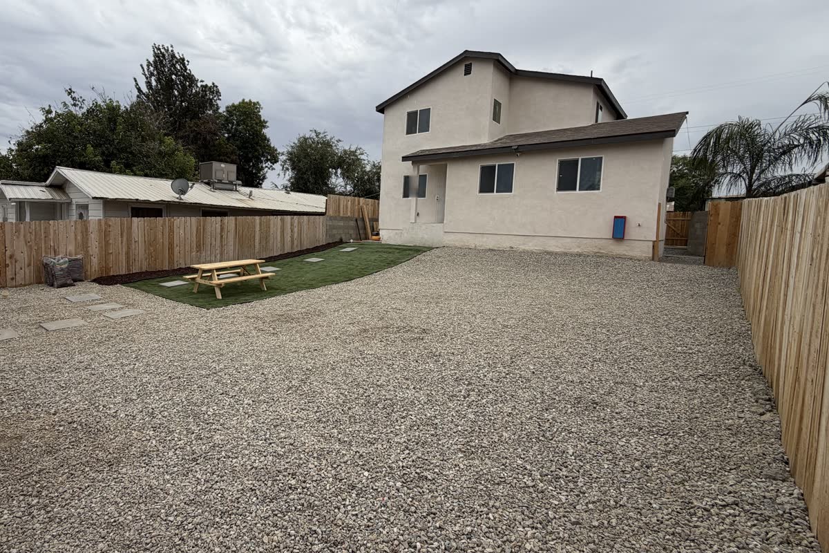 The fenced back yard seen from the far corner, gravel with a patch of lawn and a picnic table, the house at the far end